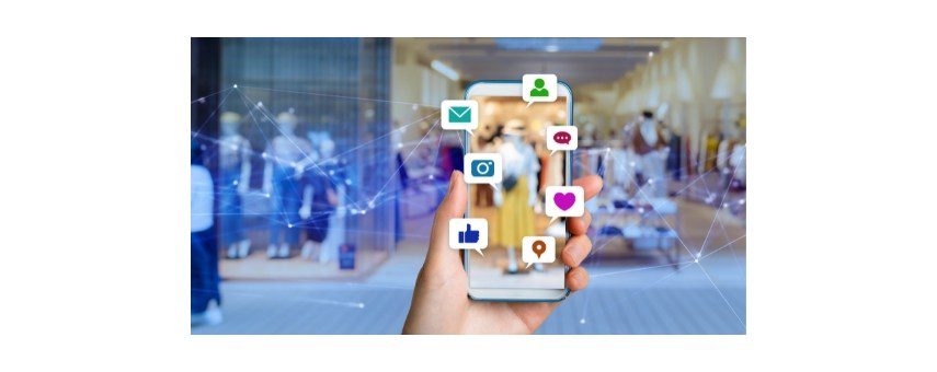 A person holds a smartphone with various app icons floating around it, including an email, camera, map pin, heart, and thumbs up. The background shows a blurred image of a clothing store interior.