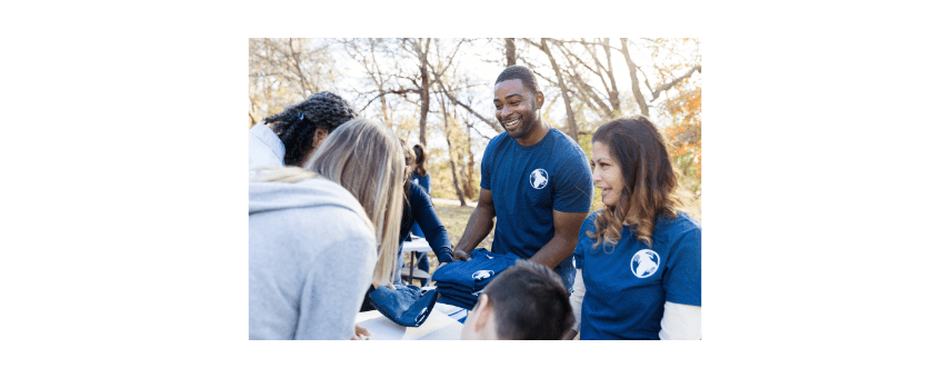 A group of people wearing matching blue shirts with a globe logo are gathered outdoors. They are smiling and engaged in conversation, with trees and a bright sky in the background.