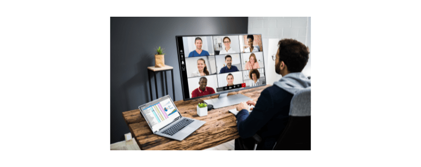 A person sits at a desk with a laptop and a large monitor displaying a video conference with multiple participants. The desk has a small plant and a coffee cup. The room has a modern, minimalist design.