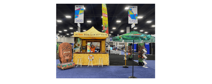 A tiki-style booth at a trade show features a thatched roof, colorful decorations, and a wooden tiki statue. An umbrella shades the area, with a person inside the booth. Banners with numbers 5600 and 5700 are visible above.