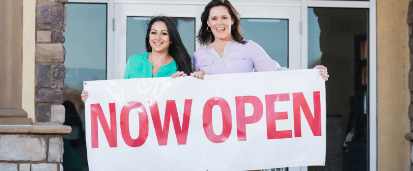 Two smiling women stand in front of a building entrance, holding a large sign that reads NOW OPEN, alongside necessary Covid signages.