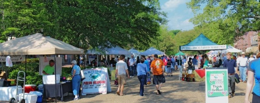 People stroll through an outdoor farmers market lined with vendor tents on a sunny day. Shoppers browse fresh produce and local goods, with trees providing shade. A sign in the foreground reads Think Green.
