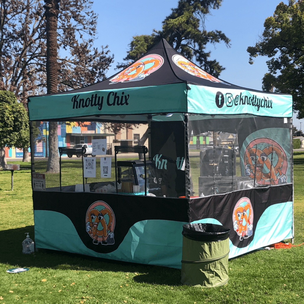 A blue and black pop-up tent labeled Knotty Chix, adorned with cartoon chicken logos, is set up in a park. It displays social media information and menu signage. A trash bin sits on the grass in front, with trees and a mural providing a picturesque backdrop.