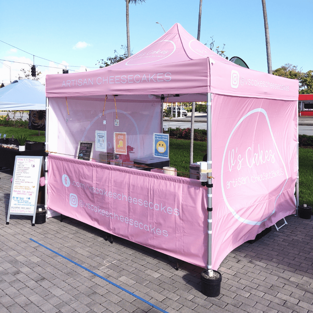 A pink pop-up tent with a canopy sells artisan cheesecakes. The booth features signage with social media handles, and a menu board is displayed on the left side. Set up on a paved area, it offers a charming view of greenery and the street in the background.