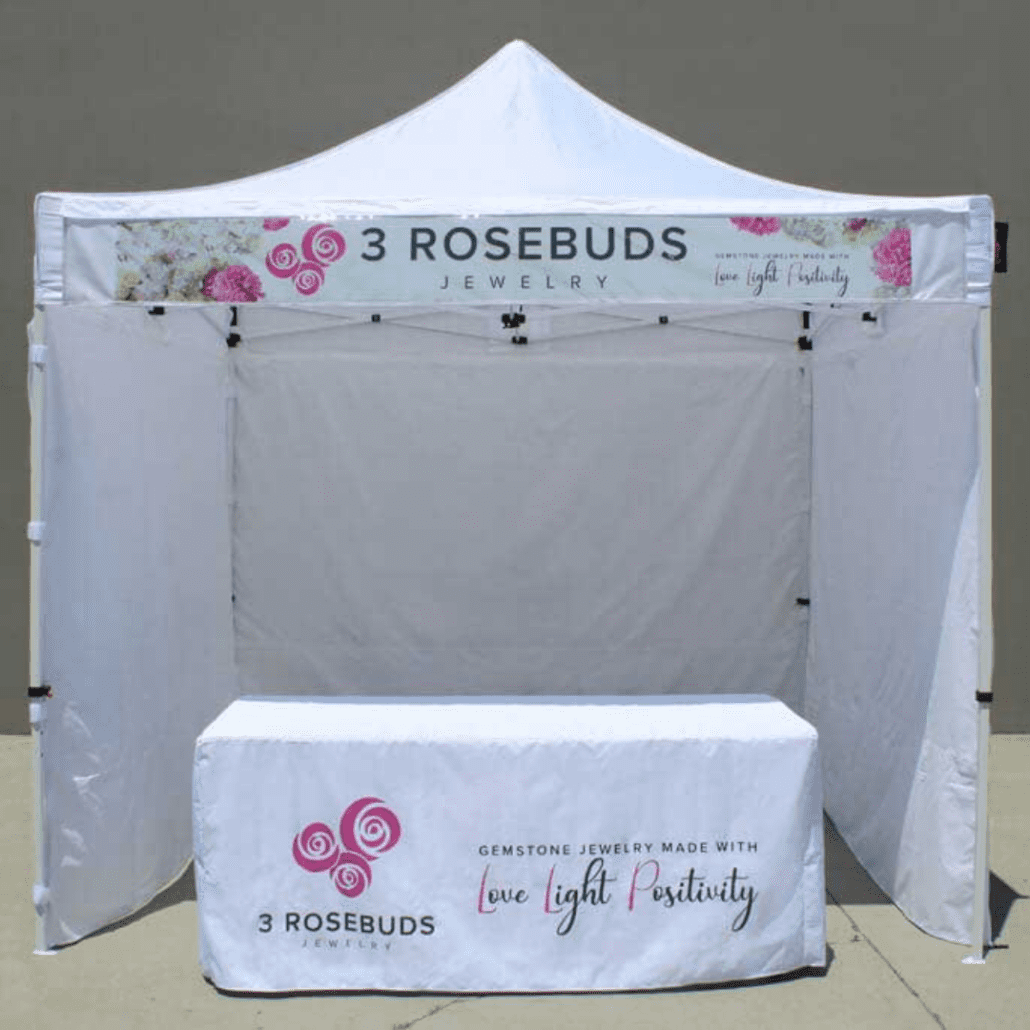 A white pop-up tent features a canopy and table adorned with the 3 Rosebuds Jewelry logo and floral graphics. The display proudly showcases the phrase "Gemstone Jewelry Made with Love, Light, Positivity," inviting passersby to explore its treasures.
