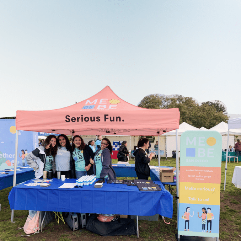 A group of people smiling under a pink pop-up tent that reads Serious Fun. They stand behind a table with pamphlets and merchandise. A sign next to them displays ME BE; San Diego, inviting inquiries about services at this lively outdoor event.