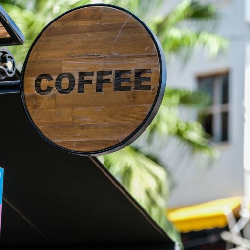 Wooden sign with the word COFFEE in bold letters, attached to the exterior of a building. The background features blurred greenery and a building with a window.
