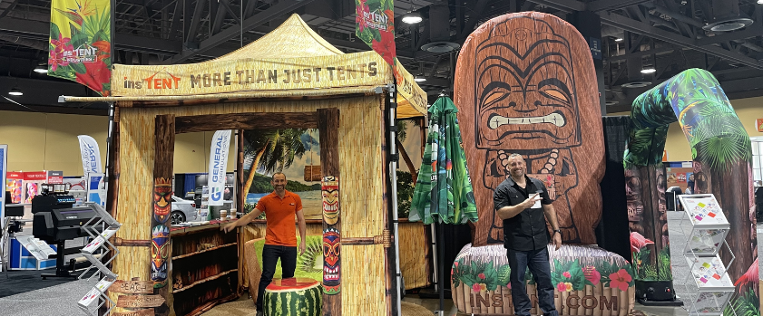 Two people pose in a tropical-themed booth at the trade show. The setup features tiki decorations, a bamboo counter, and a large tiki face structure. Signs read More Than Just Tents and InstaTent, creating a bright and colorful display with vivid tropical motifs.