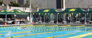 The outdoor swimming competition features participants racing in a pool with lane markers, while spectators and officials gather under custom tents marked Golden West Swim Club. In the backdrop, buildings and trees frame the lively scene.