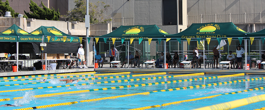 The outdoor swimming competition features participants racing in a pool with lane markers, while spectators and officials gather under custom tents marked Golden West Swim Club. In the backdrop, buildings and trees frame the lively scene.