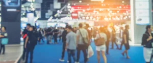 A busy trade show with people walking on a blue carpeted floor. Stalls and booths are visible with bright, colorful lights creating a lively atmosphere. The image is slightly blurred, conveying a sense of movement and activity.
