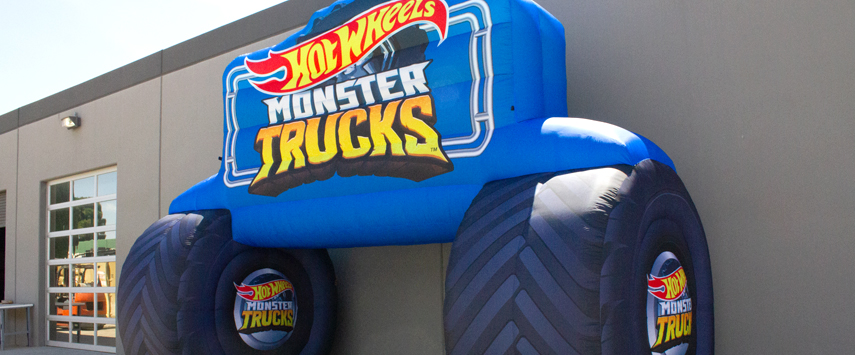 A large inflatable structure, an impressive replica of a monster truck with Hot Wheels Monster Trucks branding, is set up outside a gray building. The structure boasts big black wheels and a colorful logo on the side.