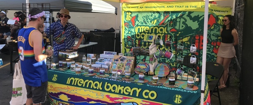A colorful market stall for Internal Bakery Co features vibrant artwork, jars, and various items at the festival exhibit. A person behind the table talks to another visitor, while a woman stands to the side. The atmosphere is bright and lively.