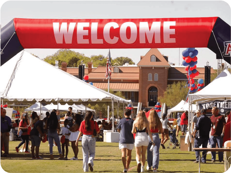 People walk under a large red WELCOME arch featuring inflatables at an outdoor event. Tents, adorned with banners and balloons, create a festive atmosphere. An American flag waves proudly, while a building with a tower looms in the distance, adding to the event's charm.