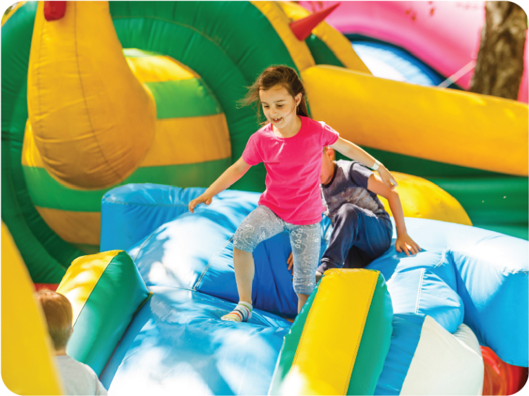 A young girl wearing a pink shirt and patterned leggings plays on a colorful inflatable bouncy castle, as highlighted in the inflatables essential guide. She climbs over a blue section, with other children and vibrant inflatable structures bustling in the background.