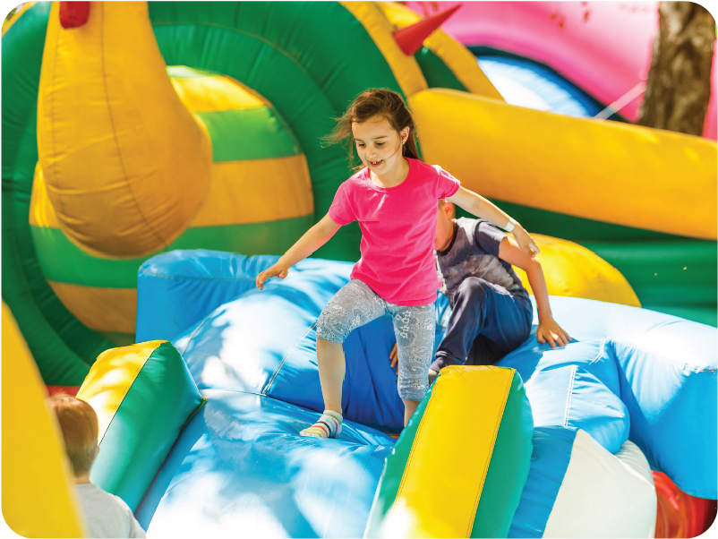 A young girl wearing a pink shirt and patterned leggings plays on a colorful inflatable bouncy castle, as highlighted in the inflatables essential guide. She climbs over a blue section, with other children and vibrant inflatable structures bustling in the background.