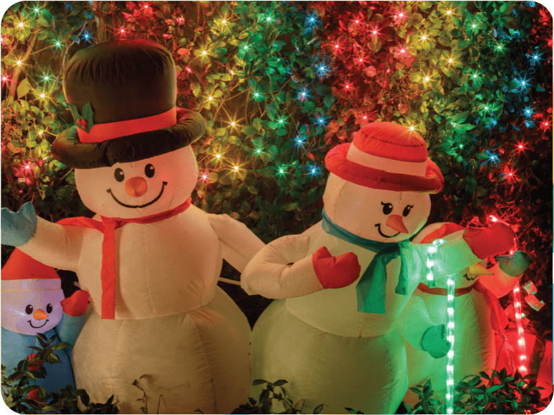 Inflatable snowmen with red and green scarves are illuminated by vibrant multicolored Christmas lights, creating a festive holiday scene. A smaller snowman peeks from the side amidst glowing foliage, showcasing an inflatables hub that's perfect for any winter wonderland.