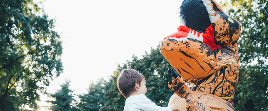 A child reaches up playfully towards someone dressed in one of those whimsical inflatable costumes, this one an enormous dinosaur. They are outdoors, surrounded by trees under a clear sky.