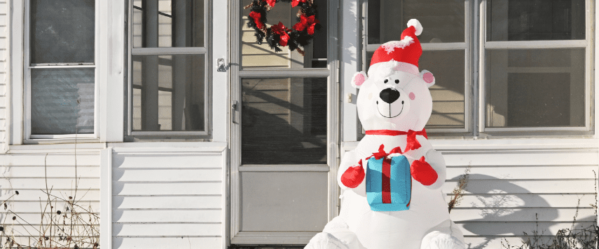 An adorable inflatable polar bear dressed in a Santa hat and scarf, holding a blue gift box, sits cheerfully on the snow-covered porch. This charming Christmas inflatable perfectly complements the white house's front door adorned with a festive red and green wreath.