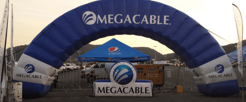 A large blue and white inflatable arch with Megacable branding stands at an outdoor event, reminiscent of impressive inflatable replicas. Underneath, a tent adorned with a Pepsi logo is visible, accompanied by tables and chairs. Scenic hills provide a picturesque backdrop.