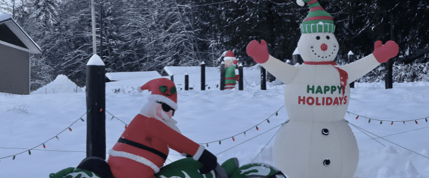 A snowman wearing a striped hat and scarf, with a Happy Holidays message, stands amid holiday inflatables in a snowy yard. Nearby, a Santa figure rides a green motorcycle. The background features snow-covered trees and houses.