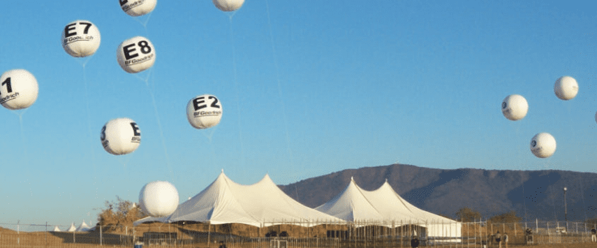 White tents are set up in an open area beneath a clear blue sky. Large inflatable signs with black text, including E1, E2, and E7, float above. A mountain range graces the background.