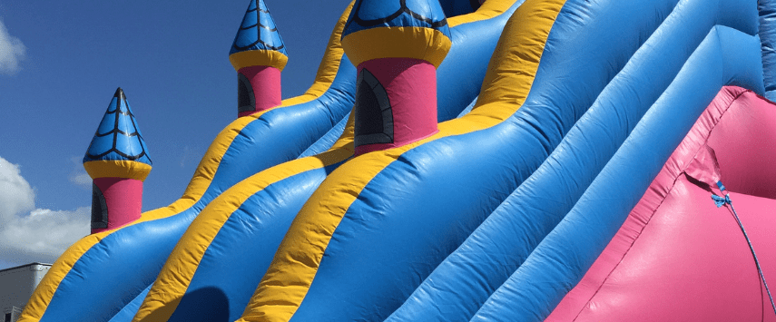 The bright inflatable slide in pink, blue, and yellow is shaped like a whimsical castle with pointed roofs. Set against a clear blue sky, this delightful inflatable slide features three distinct lanes for sliding down.