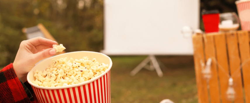 A person holds a red and white striped bucket of popcorn in an outdoor setting. An inflatable movie screen looms in the background, casting a soft glow over a wooden table with a red cup on it, suggesting a cozy outdoor movie night.