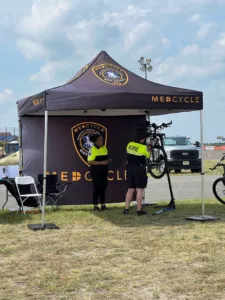 Two people wearing nurse uniforms work under a MedCycle tent. One person stands while the other repairs a bicycle on a stand. A white chair and a vehicle are visible in the background. The sky is cloudy.