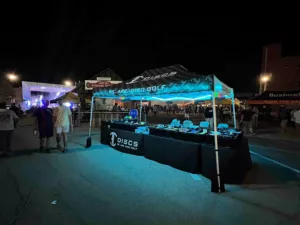 A nighttime outdoor event with a brightly lit tent displaying various discs on a table. People walk nearby under a dark sky, with a stage and buildings visible in the background.