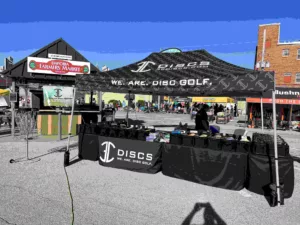 A disc golf booth at an outdoor market features a black tent with We Are Disc Golf printed on it. Discs are displayed on tables. The Emporia Farmers Market sign is visible in the background under clear skies.