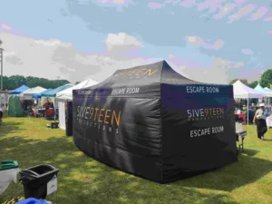 A black tent at an outdoor event labeled Five9teen Productions Escape Room with other similar tents in the background on a grassy field, under a partly cloudy sky. People are visible around the tents.
