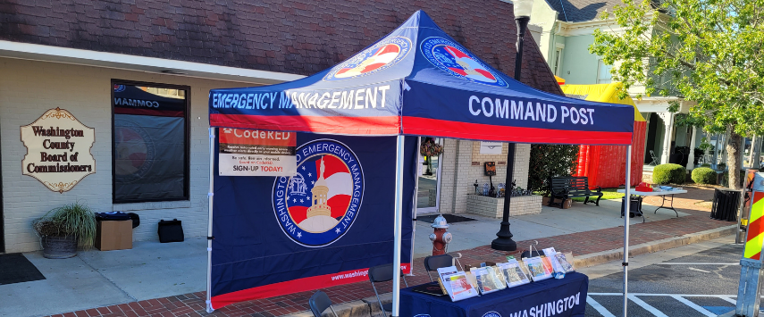 A pop-up tent labeled Emergency Management Command Post is set up outside a building with a sign saying Washington County Board of Commissioners. The pop-up tent has informational brochures on a small table and chairs nearby, ready for quick deployment and efficient crisis coordination.