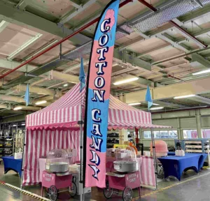 A pink and white striped tent is set up indoors with two pink cotton candy machines in front. A tall banner reads COTTON CANDY. The setup is in a spacious room with exposed pipes and industrial lighting.