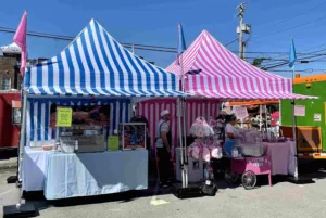 A vibrant outdoor scene features two striped tents: one blue and white with baked goods, and one pink and white with cotton candy. People are interacting with vendors beneath sunny skies, creating a festive atmosphere.