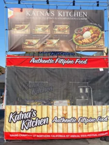 Outdoor food stall for Katnas Kitchen, offering authentic Filipino cuisine. Features a large banner and smaller signs advertising dishes like BBQ and Lechon Kawali, with the title of Grand Champion of Northern California Sisig Festival displayed.