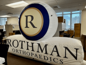 An inflatable display features a large circle with the letter R and the words Rothman Orthopaedics underneath. The display is set up in an office with boxes and desks visible in the background.