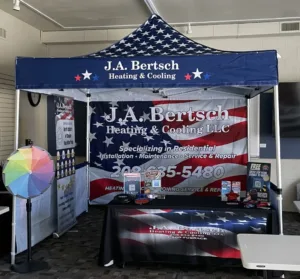 A promotional booth for J.A. Bertsch Heating & Cooling LLC, featuring an American flag design. The setup includes a canopy, display tables with brochures, a phone number, and a colorful spinning wheel on the left.