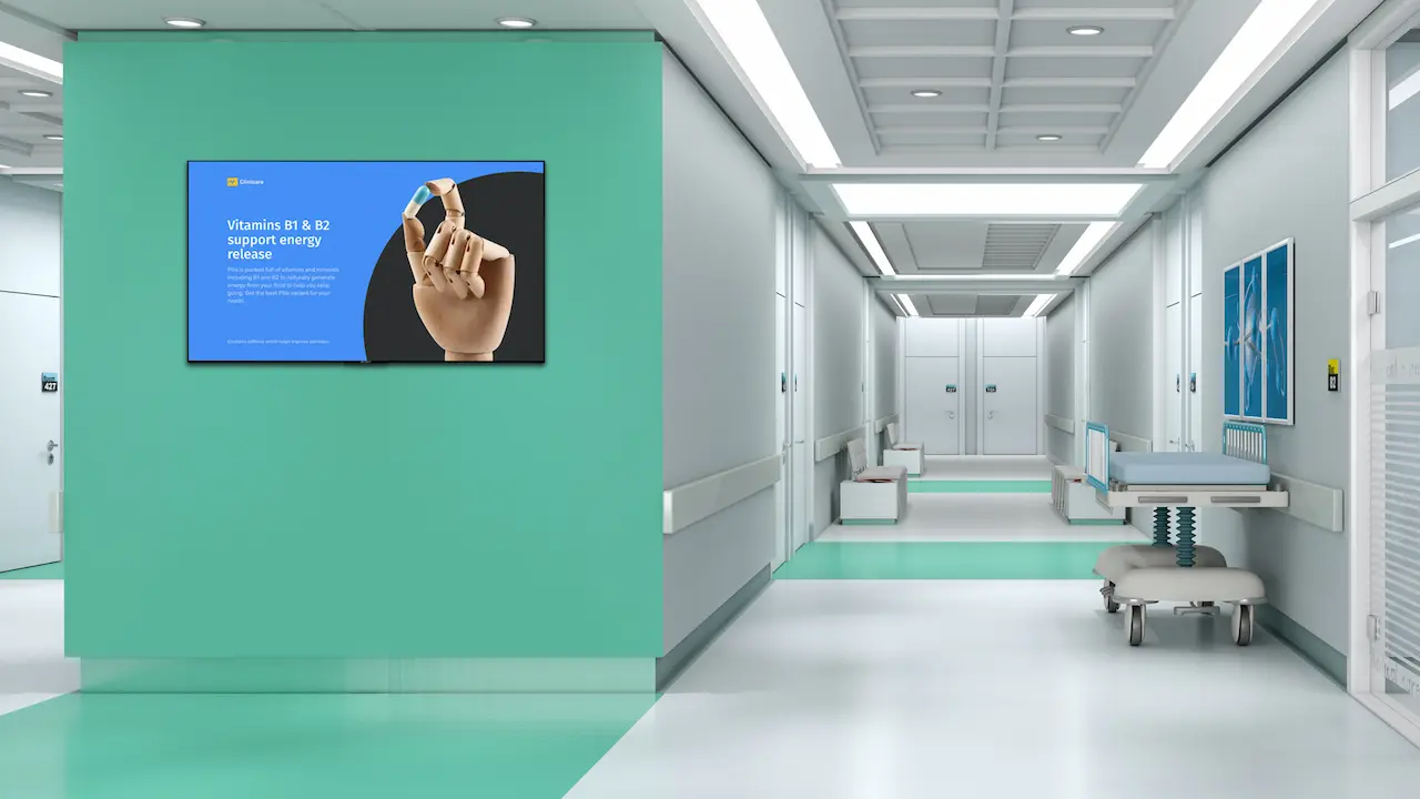 A modern hospital corridor with a mint green and white color scheme features a custom display on the wall. The digital screen shows a hand holding a small capsule, advertising vitamins B1 and B12. Two hospital beds are partially visible along the corridor.
