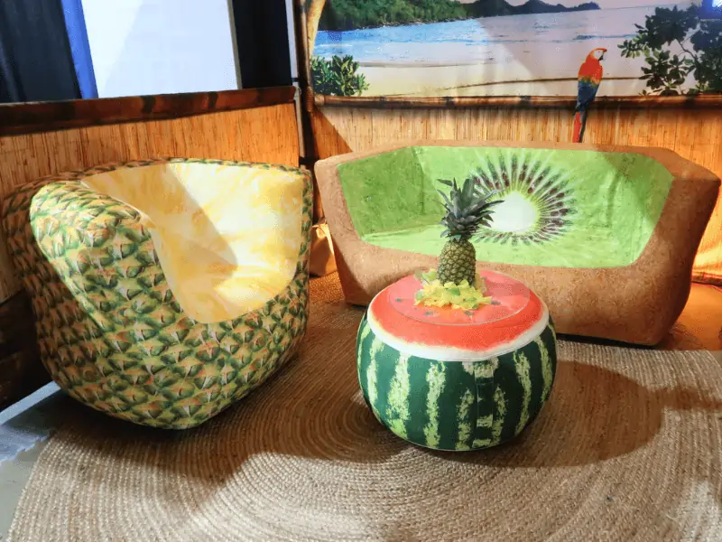 A cozy seating area features a pineapple-patterned armchair, an inflatable kiwi-themed sofa, and a watermelon ottoman. A tropical backdrop and a straw rug complete the fruity decor.