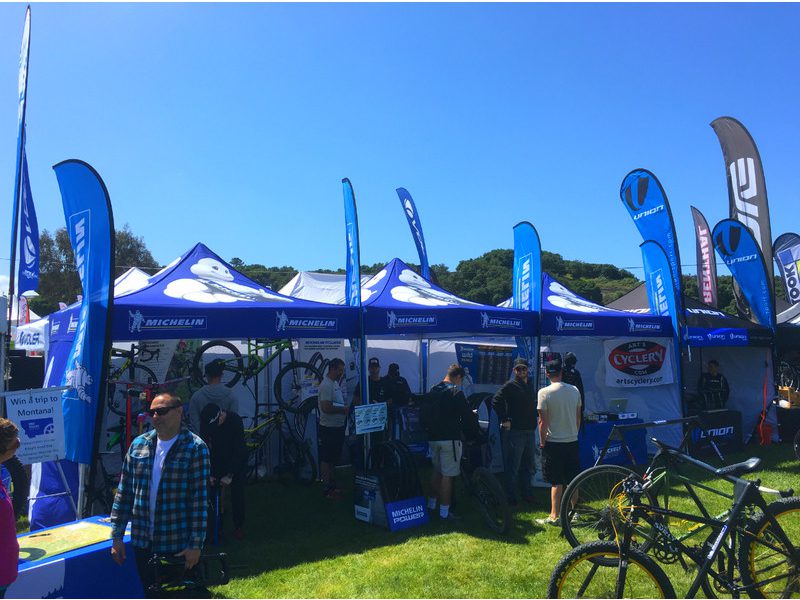 A group of people gather around a booth at an outdoor event featuring 10'x10' Custom Printed Pop Up Tents with Michelin branding and flags. Bicycles are on display, and the scene is set on a sunny day with grassy surroundings.