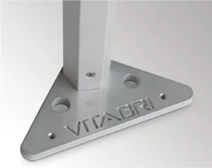 A close-up image of a metal base plate engraved with "VITABRI," designed to support the vertical column of a 10 x 10 canopy. It features three circular holes, likely intended for anchoring and securing the canopy tent in place.