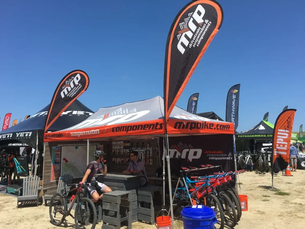 Outdoor bike event featuring a tent with MRP branding and several bicycles on display. People sit and stand around the booth under a clear blue sky. Multiple promotional flags are visible in the background.
