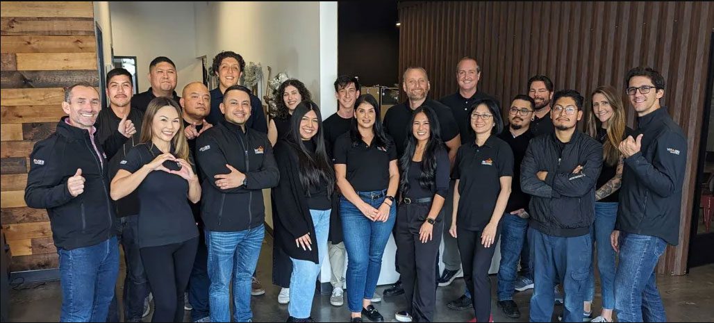 A group of 18 people smiling and posing for a photo in an indoor setting, surrounded by custom tents that add a unique flair. Clad in casual outfits, mostly in dark colors, they stand against a backdrop featuring wooden and metal elements.