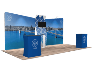 The 10'x20' Quick-N-Fit Booth - Package # 1216 showcases a large backdrop depicting a cityscape and bridge over water. It also features a central stand equipped with a screen and two smaller counters covered in blue cloth adorned with a geometric logo, all set upon wood-paneled flooring.
