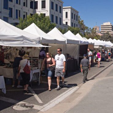 A sunny outdoor market with people walking past white tents. Vendors display items under the tents. A few shoppers browse the stalls. Visible buildings and trees in the background.