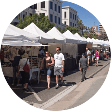People are walking past white canopy tents at an outdoor market on a sunny day. Tables under the tents display various items for sale. Buildings and trees are visible in the background.