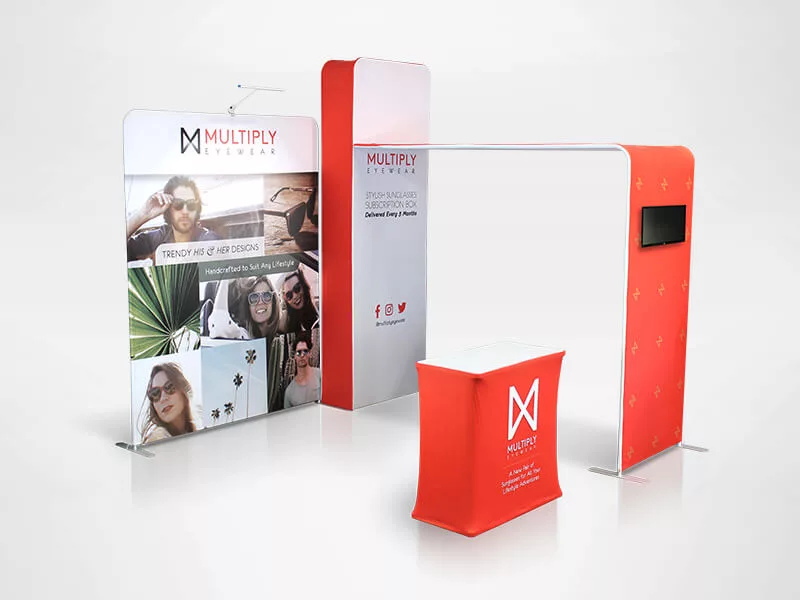 A trade show booth display with Multiply Eyewear branding, featuring images of people wearing sunglasses. The setup includes a tall backdrop, a side panel with a screen, and a small covered table, all in white and red with logos and social media icons.