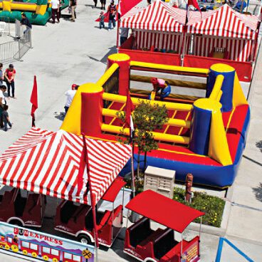 A colorful carnival scene featuring red and white striped tents and an inflatable obstacle course in vibrant blue, red, and yellow. A small red and white train is parked nearby. A few people are enjoying the activities.