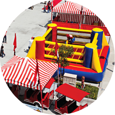 A brightly colored inflatable obstacle course in an outdoor area, surrounded by red and white striped tents. People are walking around, and a small train is visible in the foreground.
